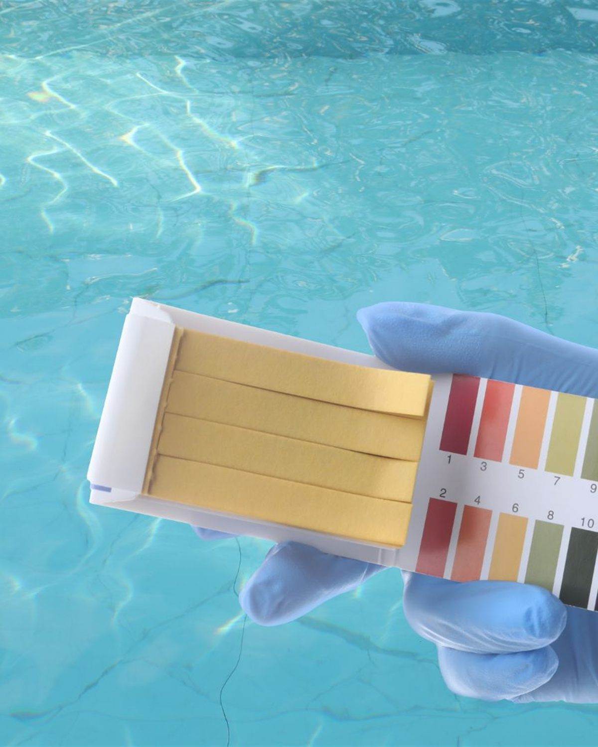 Gloved hand holding a pH test strip chart over turquoise pool water